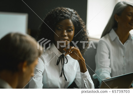 Diverse team brainstorming in office. Highlighted focus on African-American young woman sitting at table along with grey-haired mature woman and Caucasian man 65739703