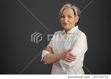 Intelligent silver-haired woman smiles with arms crossed. Pretty business woman looking at camera confidently. Isolated on gray background. Copyspace at left side. Success concept 65739710