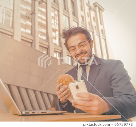 Businessman eats in fast food restaurant typing on smart phone. Young man has a break sitting at table outdoors against background of beautiful city building. Business on the go concept. Toned image 65739778