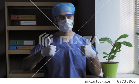 Doctor shows Thumbs up gesture. Positive gesture from male medic standing near the window against the background of a medical office. Pandemic ending concept Doctor shows Thumbs up gesture. Positive gesture from male medic standing near the window against the background of a medical office. Pandemic ending concept 65739866