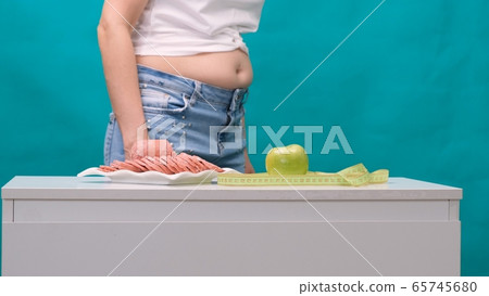 worried woman with a fat belly should choose between a green apple and a sausage. Dieting concept worried woman with a fat belly should choose between a green apple and a sausage. Dieting concept 65745680