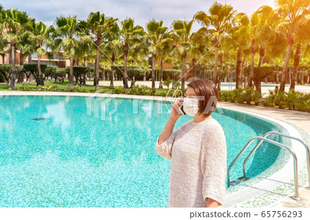 A middle-aged woman, on a sunny day, during the epidemic of the coronavirus, in a mask, talking on the phone, near the pool, in the hotel. 65756293