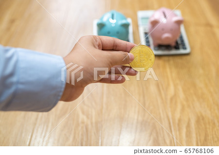Businesswoman hand holding bitcoin with piggybanks and calculator behind 65768106
