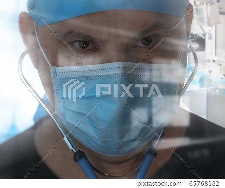 Portrait tired doctor with brown eyes in medical face mask looking at camera through window Portrait tired doctor with brown eyes in medical face mask looking at camera through window 65768182
