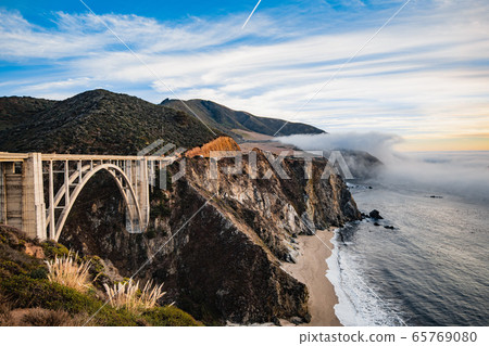 Bixby bridge foggy sunset landscape in Big Sur area on the California coast with ocean waters crashing on the shore. 65769080