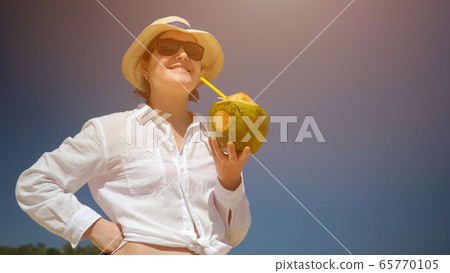 lady stands and drinks coconut with straw against blue sky 65770105