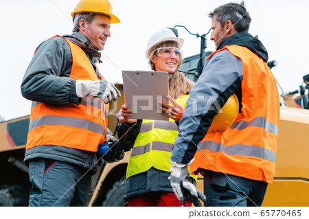 Three workers in a quarry discussing in front of heavy machinery 65770465