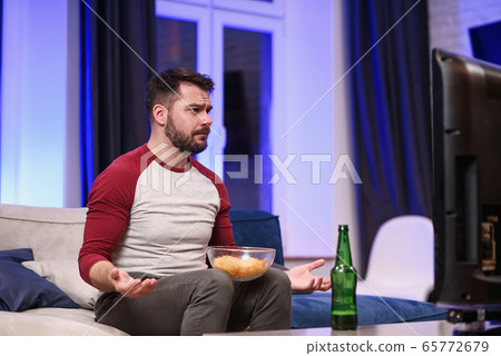 Good-looking modern positive 30-aged guy with well-groomed beard savoring chipps during emotional review of favourite football team's game on tv at home 65772679