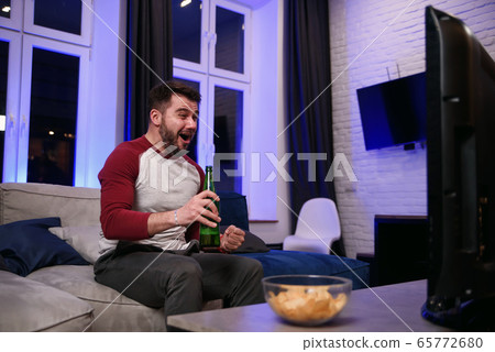 Good-looking modern positive 30-aged guy with well-groomed beard savoring chipps during emotional review of favourite football team's game on tv at home 65772680