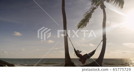 Woman lies in hammock wearing summer dress and hat watching sunset with ocean in the distance Woman lies in hammock wearing summer dress and hat watching sunset with ocean in the distance 65775729