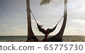 Woman lies in hammock wearing summer dress and hat watching sunset with ocean in the distance 65775730