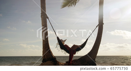 Woman lies in hammock wearing summer dress and hat watching sunset with ocean in the distance Woman lies in hammock wearing summer dress and hat watching sunset with ocean in the distance 65775730