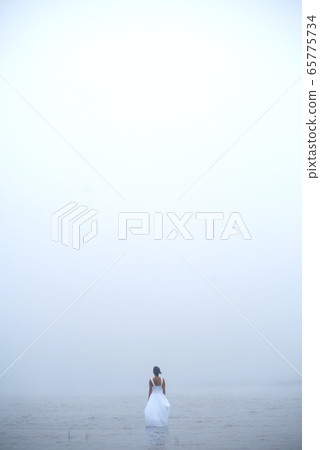 Woman in white dress and blue hair looks as if she is drifting by water surrounded by mist and fog  65775734