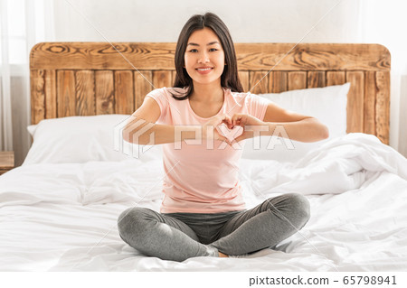 Asian Woman Gesturing Heart Shape Sitting On Bed At Home 65798941