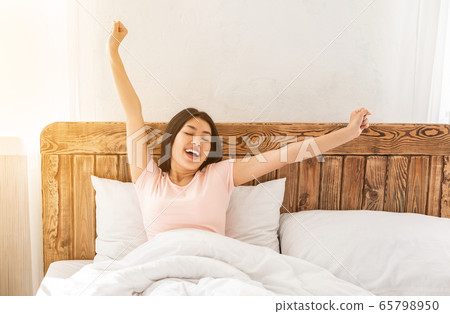 Happy asian woman yawning waking up in bed at home Happy asian woman yawning waking up in bed at home 65798950