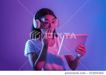 Surprised asian girl in big headphones closes her mouth with hand and looks at tablet Surprised asian girl in big headphones closes her mouth with hand and looks at tablet 65800488