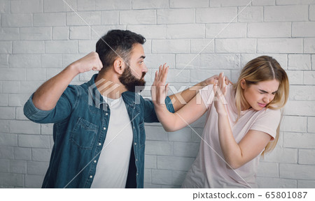 Angry man brandishing fist and holding woman, frightened woman hides from blows 65801087