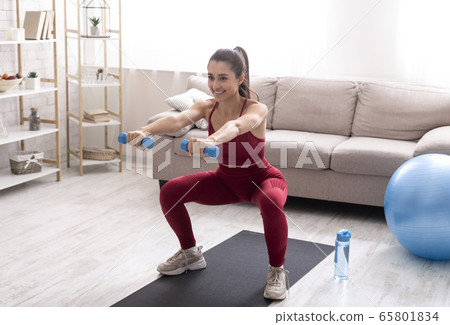 Indoor sports. Strong young woman working out with dumbbells at home, doing squats exercise 65801834