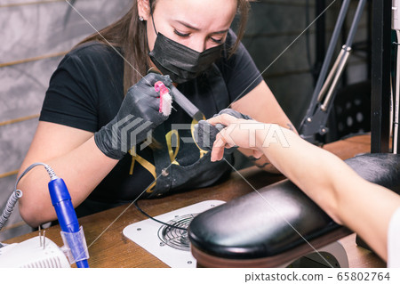 Female master uses an electric machine to remove the nail polish during manicure in the salon. Close-up hardware manicure. Concept of hand care. Female manicurist cleaning of nails by a milling cutter Female master uses an electric machine to remove the nail polish during manicure in the salon. Close-up hardware manicure. Concept of hand care. Female manicurist cleaning of nails by a milling cutter 65802764