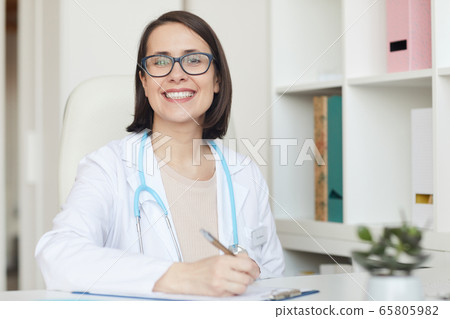 Smiling Female Doctor Posing at Desk in Clinic Smiling Female Doctor Posing at Desk in Clinic 65805982