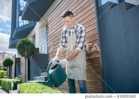Delighted dark-haired male in protective gloves and apron watering bushes with watering can, smiling Delighted dark-haired male in protective gloves and apron watering bushes with watering can, smiling 65809368