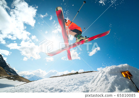 A young athlete skier does a trick on a snow kicker jump and cross skis in a counter light against a blue sky and clouds in the mountains. 65810085