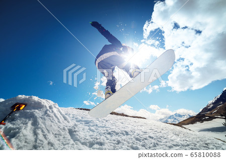 Stylish young girl snowboarder does the trick in jumping from a snow kicker against the blue sky clouds and mountains in the spring. 65810088