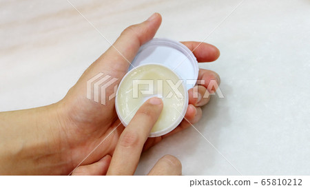 Hand holding a small round container with ointment, and a finger trying to scoop some ointment out. 65810212