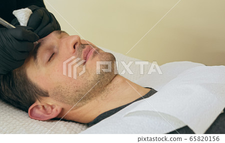 Close up of cosmetologist making permanent makeup. Microblading procedure. High quality close-up of a man laying on a couch during the esthetician brow microblading treatment. Close up of cosmetologist making permanent makeup. Microblading procedure. High quality close-up of a man laying on a couch during the esthetician brow microblading treatment. 65820156
