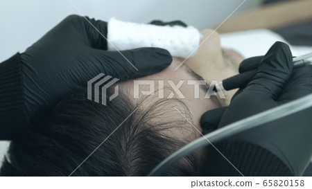 Close up of cosmetologist making permanent makeup. Microblading procedure. High quality close-up of a man laying on a couch during the esthetician brow microblading treatment. Close up of cosmetologist making permanent makeup. Microblading procedure. High quality close-up of a man laying on a couch during the esthetician brow microblading treatment. 65820158