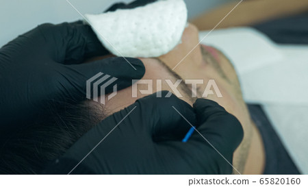 Close up of cosmetologist making permanent makeup. Microblading procedure. High quality close-up of a man laying on a couch during the esthetician brow microblading treatment. Close up of cosmetologist making permanent makeup. Microblading procedure. High quality close-up of a man laying on a couch during the esthetician brow microblading treatment. 65820160