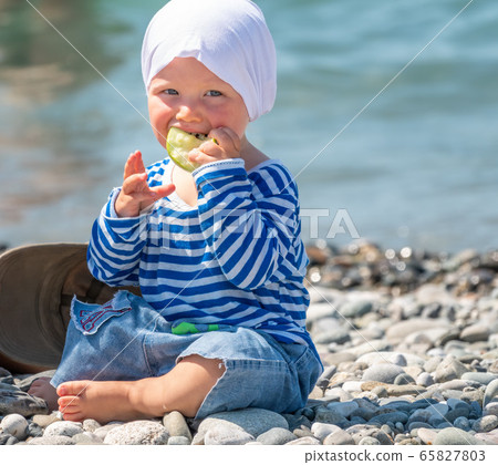 Happy one-year-old child sits on pebbles on the sea beach and eats an apple Happy one-year-old child sits on pebbles on the sea beach and eats an apple 65827803