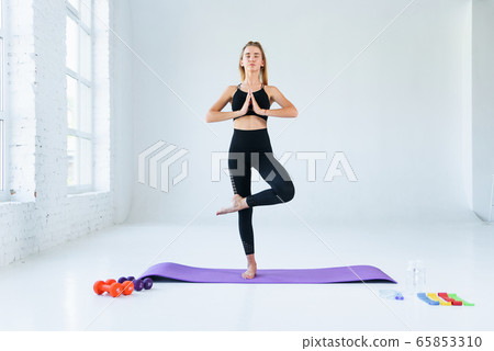 Girl practicing yoga, standing in vrksasana exercise with namaste, tree pose. Female meditation at white loft studio background. Girl practicing yoga, standing in vrksasana exercise with namaste, tree pose. Female meditation at white loft studio background. 65853310
