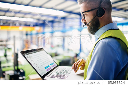 Side view of technician or engineer with headset and laptop standing in industrial factory. Side view of technician or engineer with headset and laptop standing in industrial factory. 65865824