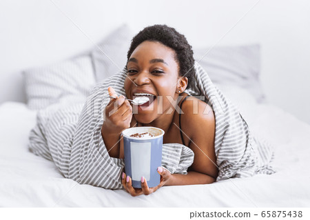 Happy african american girl puts spoon in her mouth and holds bucket of ice cream 65875438