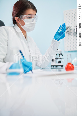 Young woman lab technician with test tube in hand makes note. Young woman lab technician with test tube in hand makes note. 65878112