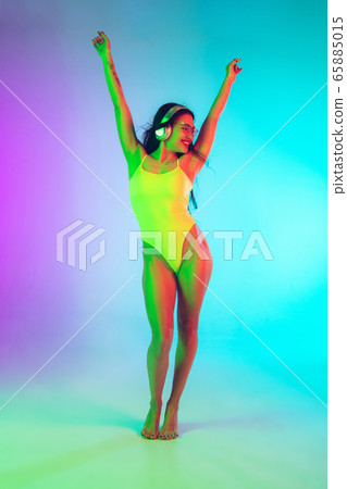 Beautiful girl in fashionable swimsuit isolated on gradient studio background in neon light. Summer, resort, fashion and weekend concept Beautiful girl in fashionable swimsuit isolated on gradient studio background in neon light. Summer, resort, fashion and weekend concept 65885015