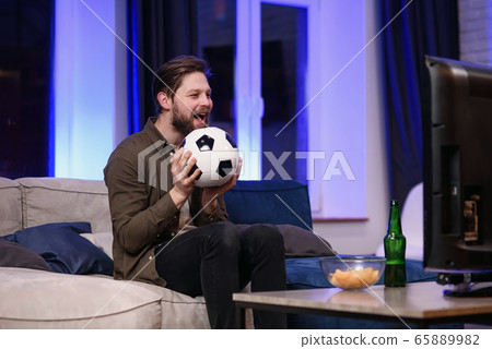 Good-looking modern positive 30-aged guy with well-groomed beard savoring chipps during emotional review of favourite football team's game on tv at home 65889982