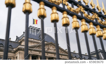 The Royal Palace in Brussels, Belgium. View through the metal fence with golden details. National flag of the Kingdom of Belgium waving on top. The Royal Palace in Brussels, Belgium. View through the metal fence with golden details. National flag of the Kingdom of Belgium waving on top. 65903670