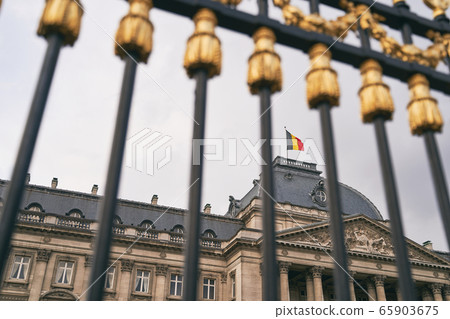 The Royal Palace in Brussels, Belgium. View through the metal fence with golden details. National flag of the Kingdom of Belgium waving on top. The Royal Palace in Brussels, Belgium. View through the metal fence with golden details. National flag of the Kingdom of Belgium waving on top. 65903675