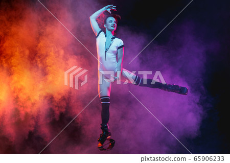 Beautiful redhead woman in a white sportswear jumping in a kangoo jumps shoes isolated on gradient studio background in neon lighted smoke 65906233