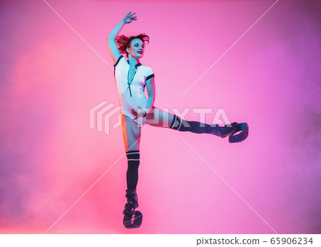 Beautiful redhead woman in a white sportswear jumping in a kangoo jumps shoes isolated on gradient studio background in neon light Beautiful redhead woman in a white sportswear jumping in a kangoo jumps shoes isolated on gradient studio background in neon light 65906234