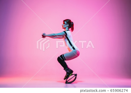 Beautiful redhead woman in a white sportswear jumping in a kangoo jumps shoes isolated on gradient studio background in neon light Beautiful redhead woman in a white sportswear jumping in a kangoo jumps shoes isolated on gradient studio background in neon light 65906274