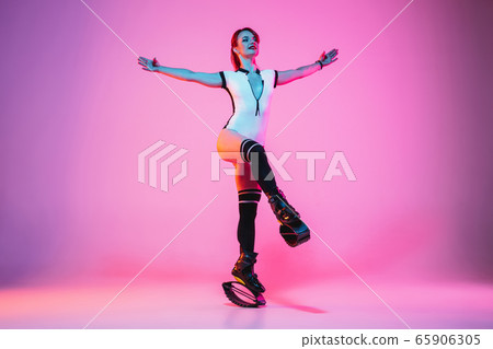 Beautiful redhead woman in a white sportswear jumping in a kangoo jumps shoes isolated on gradient studio background in neon light Beautiful redhead woman in a white sportswear jumping in a kangoo jumps shoes isolated on gradient studio background in neon light 65906305