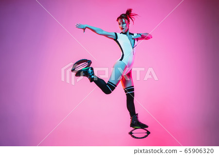 Beautiful redhead woman in a white sportswear jumping in a kangoo jumps shoes isolated on gradient studio background in neon light 65906320