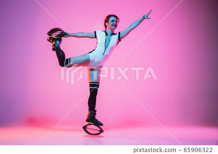 Beautiful redhead woman in a white sportswear jumping in a kangoo jumps shoes isolated on gradient studio background in neon light 65906322