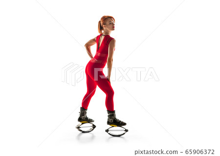 Beautiful redhead woman in a red sportswear jumping in a kangoo jumps shoes isolated on white studio background. Beautiful redhead woman in a red sportswear jumping in a kangoo jumps shoes isolated on white studio background. 65906372