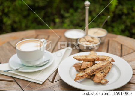 A plate of homemade biscotti cookies and a Cup of coffee with cream and sugar on a wooden table outside A plate of homemade biscotti cookies and a Cup of coffee with cream and sugar on a wooden table outside 65914909