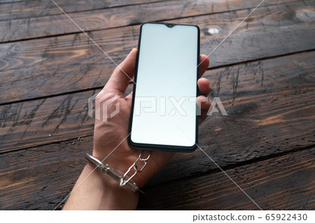 Top view men hand in handcuffs hold modern smartphone with blank screen on wooden brown background. The concept of internet and gadget dependency. Top view men hand in handcuffs hold modern smartphone with blank screen on wooden brown background. The concept of internet and gadget dependency. 65922430