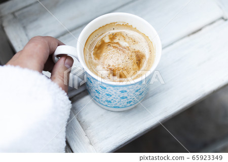 cropped female hand and cappuccino coffee in a cup on a white wooden table. Top view, copy space. cropped female hand and cappuccino coffee in a cup on a white wooden table. Top view, copy space. 65923349